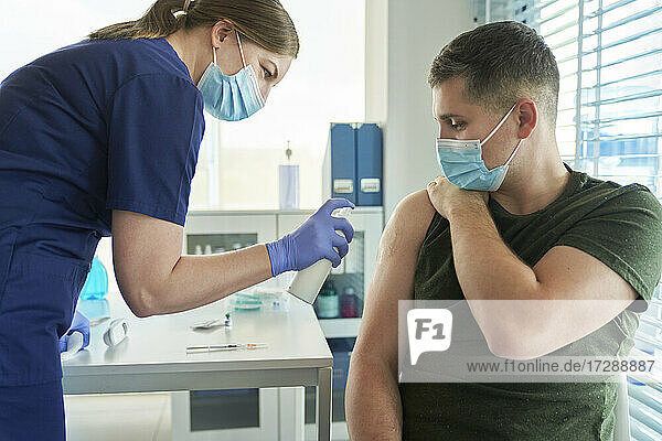 Ärztin sprüht Desinfektionsmittel auf den Arm eines männlichen Patienten  bevor sie in der Klinik den Impfstoff COVID-19 verabreicht