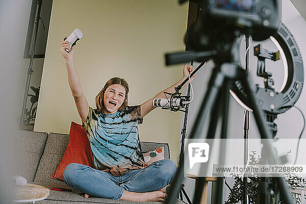 Cheerful girl with arms raised playing game while podcasting at home
