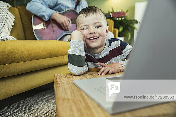 Niedlicher Junge sieht sich ein Video auf dem Laptop an  während sein Vater im Wohnzimmer Gitarre spielt