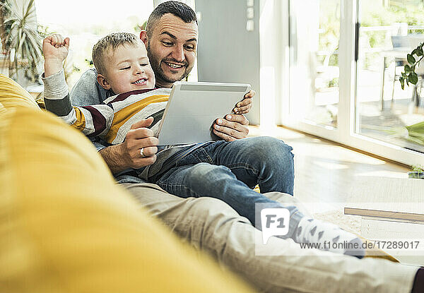 Lächelnder Vater und Sohn sehen sich ein Video über ein Tablet an  während sie auf dem Sofa im Wohnzimmer sitzen