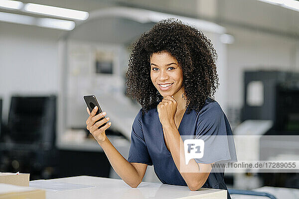 Lächelnde Geschäftsfrau mit Hand am Kinn  die ein Smartphone hält  während sie sich in der Industrie auf einen Tisch stützt