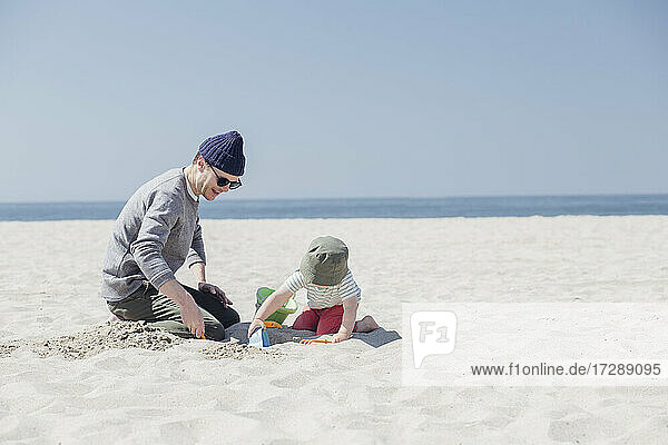 Mann und Sohn spielen mit Sandspielzeug am Strand an einem sonnigen Tag