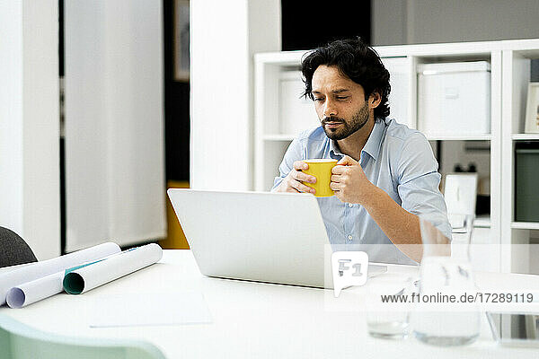 Männlicher Unternehmer mit Kaffeetasse und Laptop am Schreibtisch