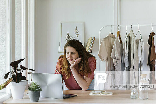 Lächelnde Geschäftsfrau mit Hand am Kinn  die zu Hause einen Laptop benutzt
