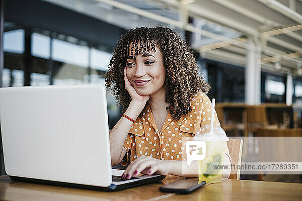Lächelnde Geschäftsfrau mit Hand am Kinn  die in einem Café einen Laptop benutzt