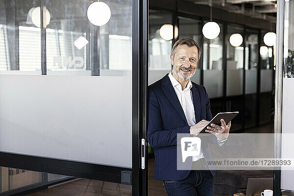 Smiling male professional holding digital tablet while leaning on glass door at office