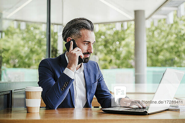 Männlicher Unternehmer  der einen Laptop benutzt  während er in einer Cafeteria mit seinem Handy telefoniert
