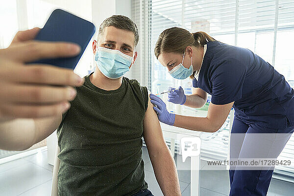 Mann mit Gesichtsmaske macht ein Selfie mit seinem Smartphone während der COVID-19-Impfung