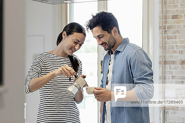 Lächelnde Frau gießt Kaffee ein  während der Mann eine Tasse in der Küche zu Hause hält