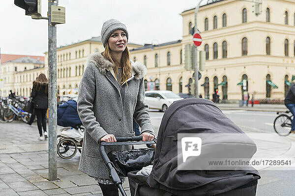 Smiling woman looking away while walking with baby stroller in city