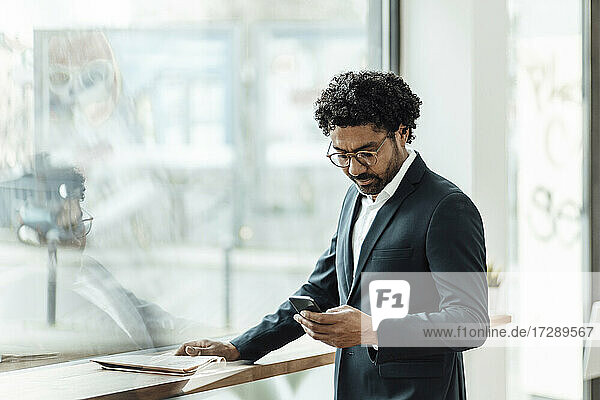 Male entrepreneur using smart phone while standing by glass window in office
