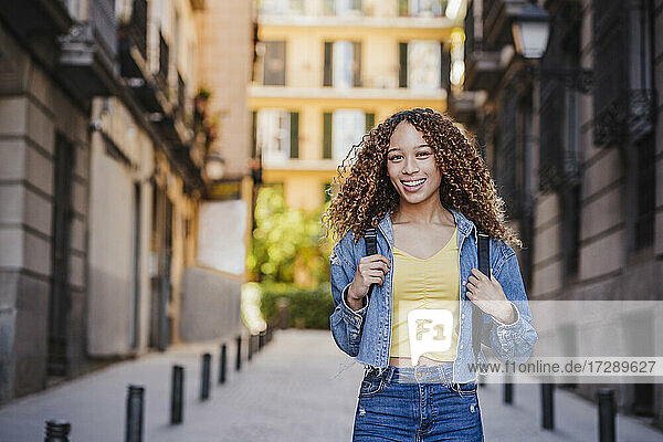 Lächelnde Frau in Jeanskleidung steht in einer Gasse in der Nähe von Gebäuden
