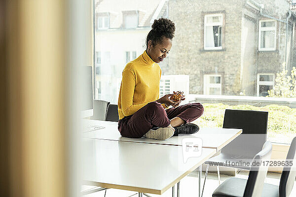 Geschäftsfrau spielt mit Puzzle-Würfel  während sie im Schneidersitz auf dem Schreibtisch im Büro sitzt