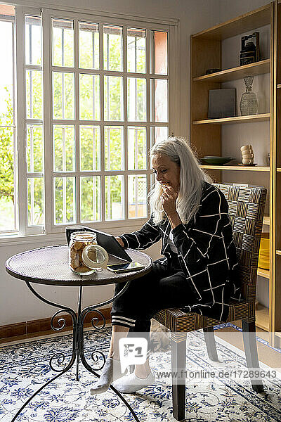 Frau isst Kekse  während sie ein digitales Tablet am Wohnzimmertisch benutzt