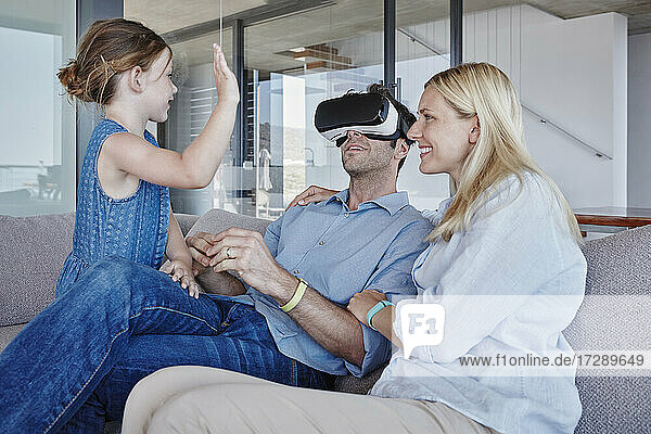 Mädchen winkt Vater zu  der einen Virtual-Reality-Simulator trägt  der bei einer Frau im Wohnzimmer sitzt