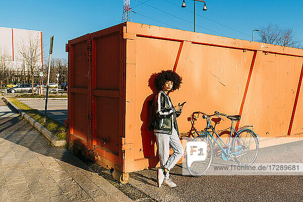 Frau mit Mobiltelefon  die sich an einem sonnigen Tag an einen Container lehnt