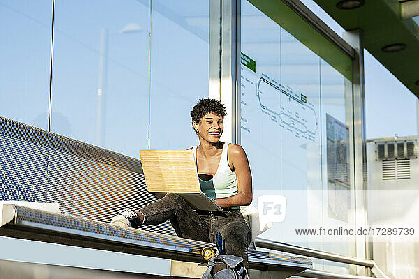 Lächelnde junge Frau sitzt mit Laptop auf einer Bank