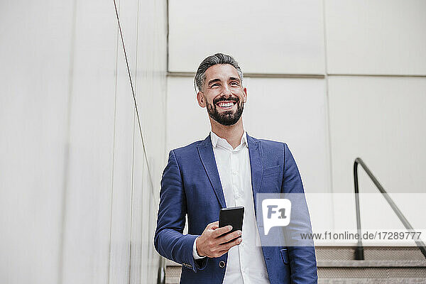 Männlicher Unternehmer mit Smartphone  der lächelnd an der Wand steht