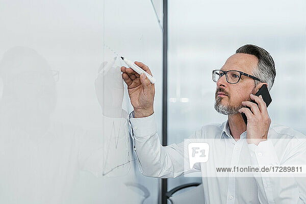 Männlicher Unternehmer  der im Büro am Whiteboard schreibt und gleichzeitig mit dem Handy telefoniert