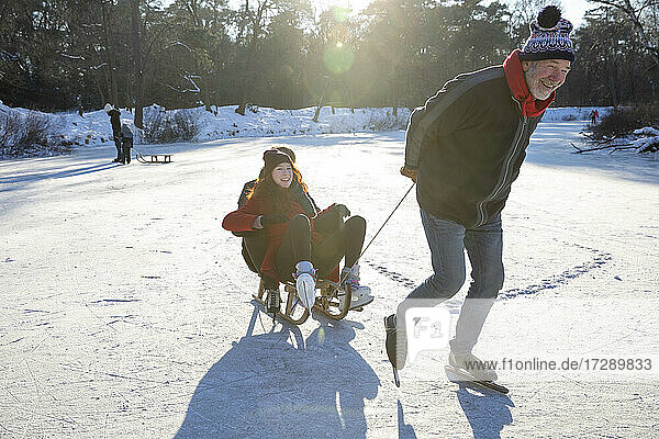 Älterer Mann mit jungem Paar  das beim Schlittschuhlaufen über einen zugefrorenen See einen Schlitten zieht