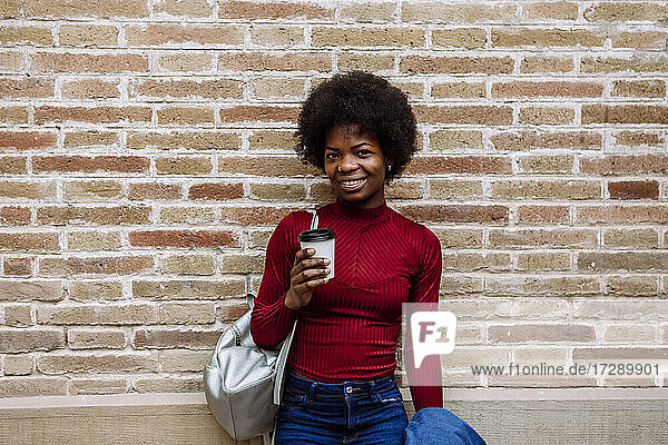 Smiling young woman with reusable cup standing in front of brick wall