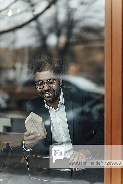 Lächelnder männlicher Unternehmer  der ein Hausmodell betrachtet  während er in einem Café sitzt  gesehen durch Glas