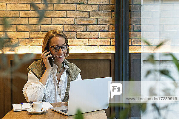 Lächelnde Frau  die einen Laptop benutzt  während sie in einem Café mit einem Mobiltelefon spricht