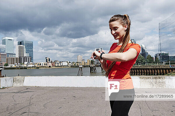Smiling fit woman checking time on wristwatch by river in city