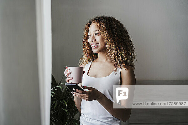 Happy woman with smart phone and coffee mug standing at bedroom