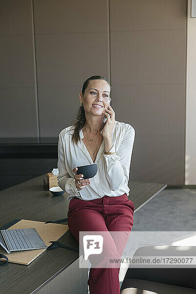 Smiling female entrepreneur holding coffee cup talking on smart phone while sitting at desk in office