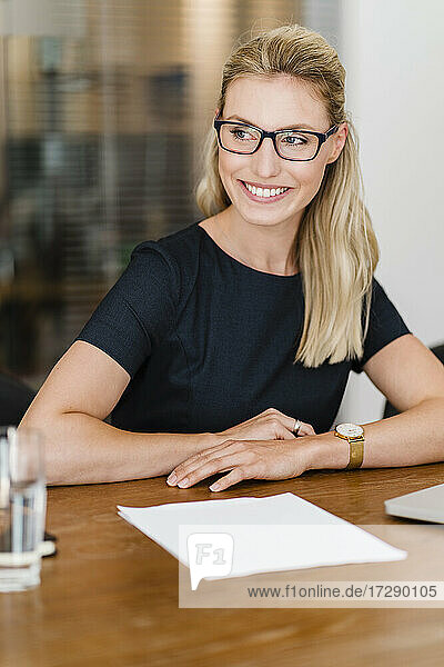 Lächelnde schöne Geschäftsfrau mit Brille  die am Schreibtisch sitzt und wegschaut