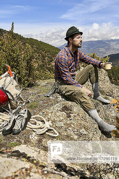 Male mountaineer holding cup while sitting on mountain during sunny day