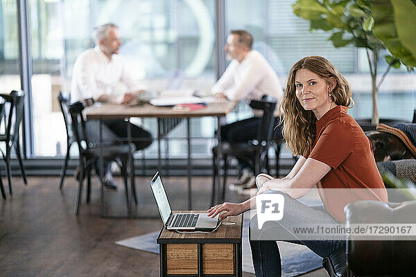 Geschäftsfrau vor einem Laptop sitzend mit männlichen Kollegen  die im Hintergrund in einer Büro-Cafeteria diskutieren