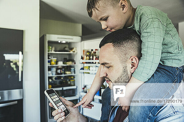 Junge sitzt auf der Schulter eines Mannes und zeigt auf sein Smartphone in der Küche