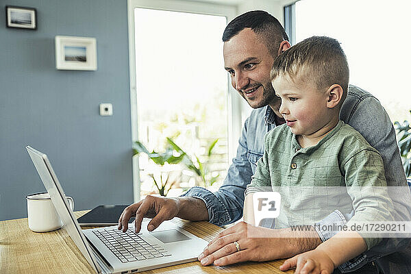 Lächelnder männlicher Freiberufler  der einen Laptop benutzt  während er mit seinem Sohn zu Hause sitzt