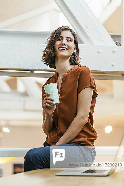 Smiling young female entrepreneur with disposable coffee cup sitting on desk at creative office