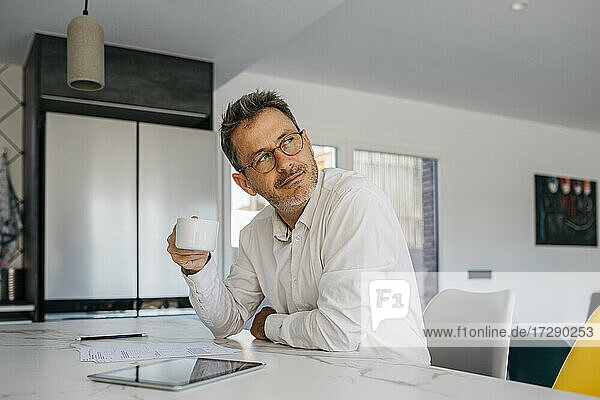 Männlicher Freiberufler mit Kaffeetasse  der wegschaut  während er in der Küche zu Hause sitzt
