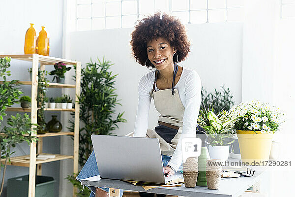 Smiling female shop owner with laptop sitting on table at plant store