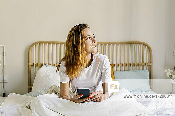 Entspannte Frau  die wegschaut  während sie ihr Handy auf dem Bett zu Hause hält