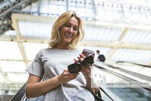 Lächelnde Frau  die ein Mobiltelefon benutzt  während sie auf einer Rolltreppe nach unten fährt