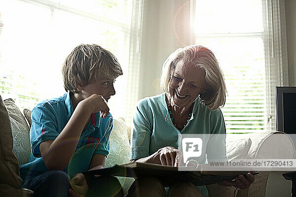 Smiling woman showing photo album to grandson while sitting on sofa at home