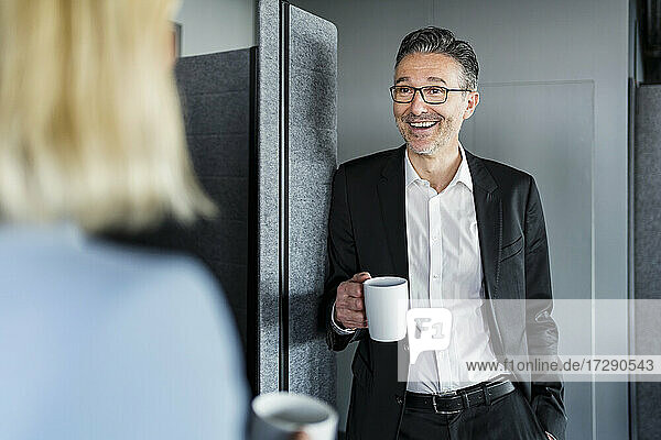 Lächelnder männlicher Unternehmer  der eine Kaffeetasse hält  während er mit einer weiblichen Fachkraft in der Pause im Büro spricht