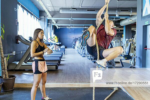 Female instructor looking at male athlete climbing rope during training at gym