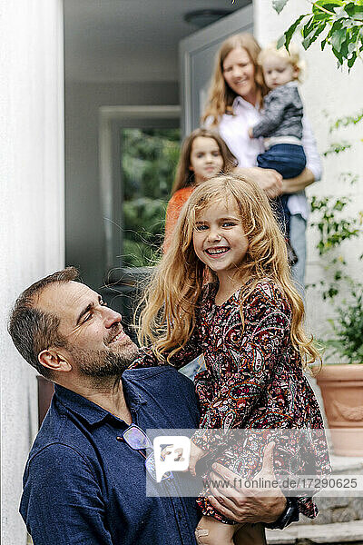 Vater trägt fröhliche Tochter  während Familie im Hintergrund außerhalb des Hauses