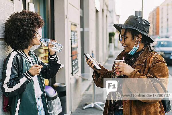 Junge Frau  die auf dem Gehweg mit einem Freund Wasser trinkt und dabei ein Mobiltelefon benutzt
