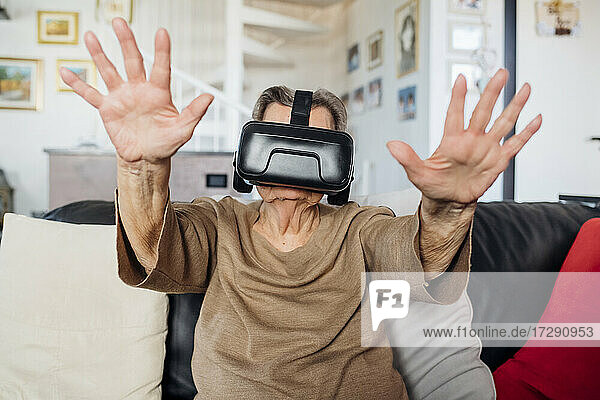 Ältere Frau trägt einen Virtual-Reality-Simulator und gestikuliert  während sie zu Hause auf dem Sofa im Wohnzimmer sitzt