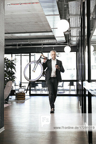 Älterer Geschäftsmann  der ein Mobiltelefon benutzt  während er im Büro ein Fahrrad auf der Schulter trägt