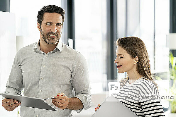 Lächelnder männlicher Fachmann  der wegschaut  während er mit einer Kollegin im Büro über ein digitales Tablet diskutiert
