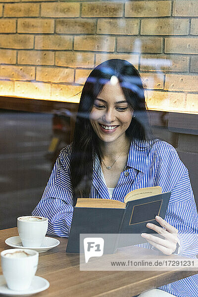 Smiling young woman reading book while sitting in bar seen through glass