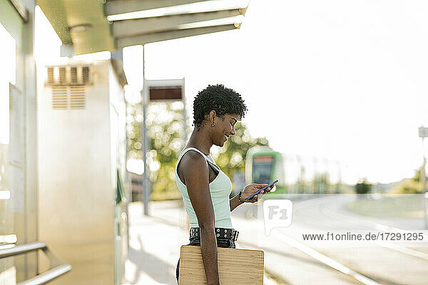 Lächelnde junge Frau mit Laptop und Smartphone an einer Straßenbahnhaltestelle